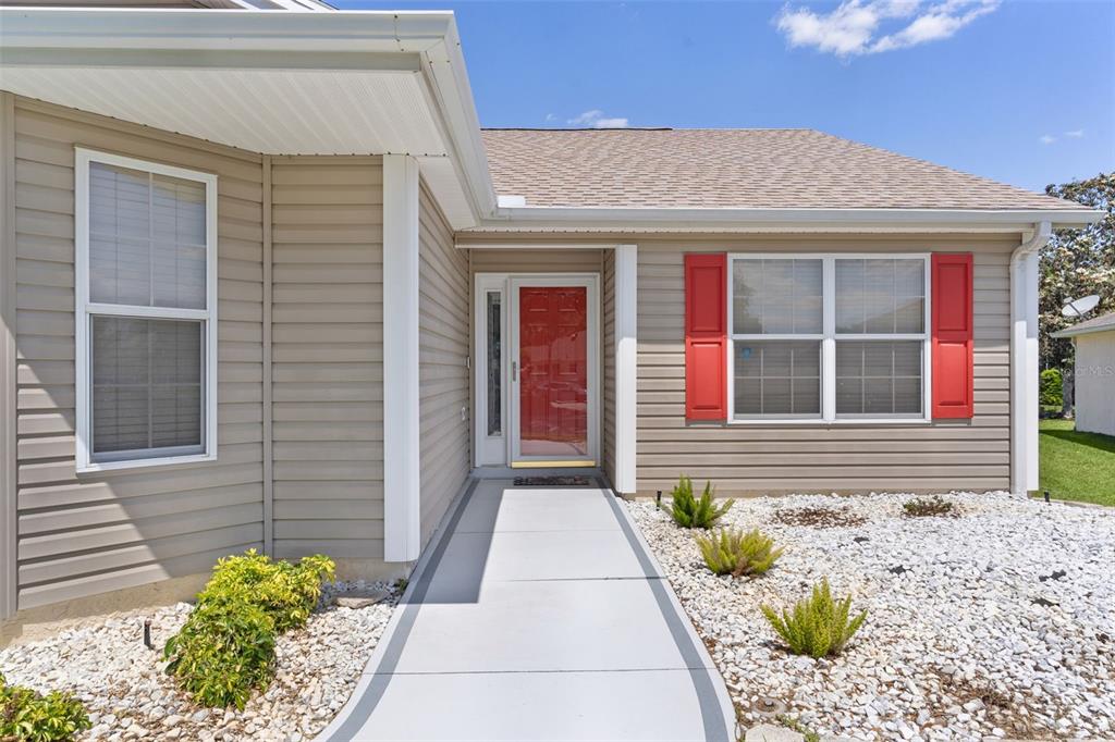 A tan-sided house features a red front door, white trim, and a concrete walkway — 17107 SE 93RD EXETER COURT, THE VILLAGES, FL