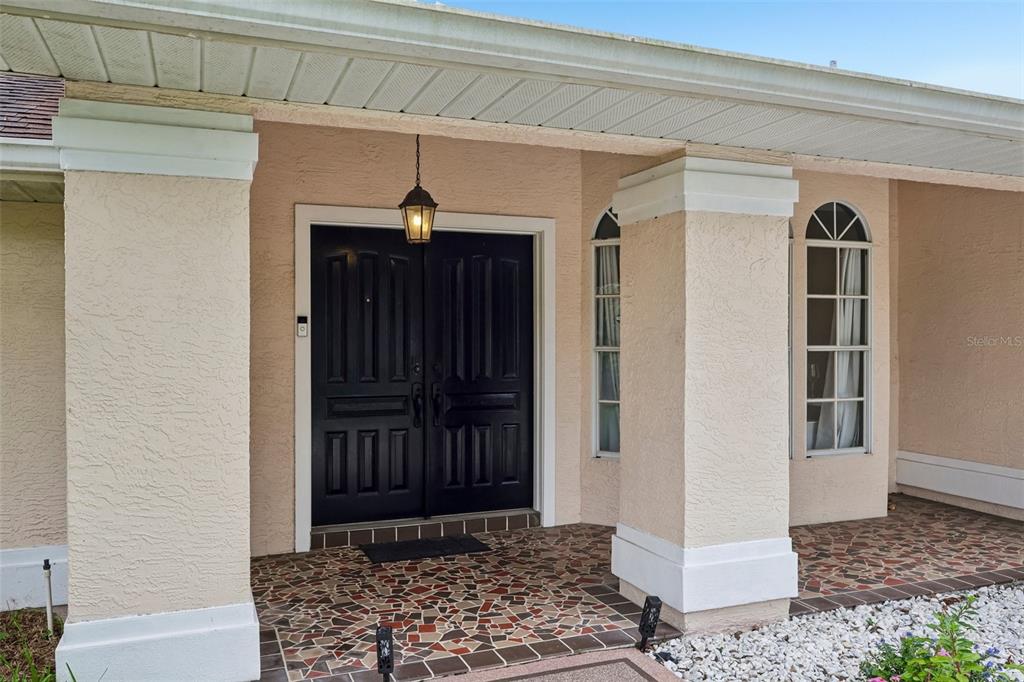 A covered front porch features a double black door, stone tile flooring, and stucco columns — 1260 MASADA LANE, SPRING HILL, FL