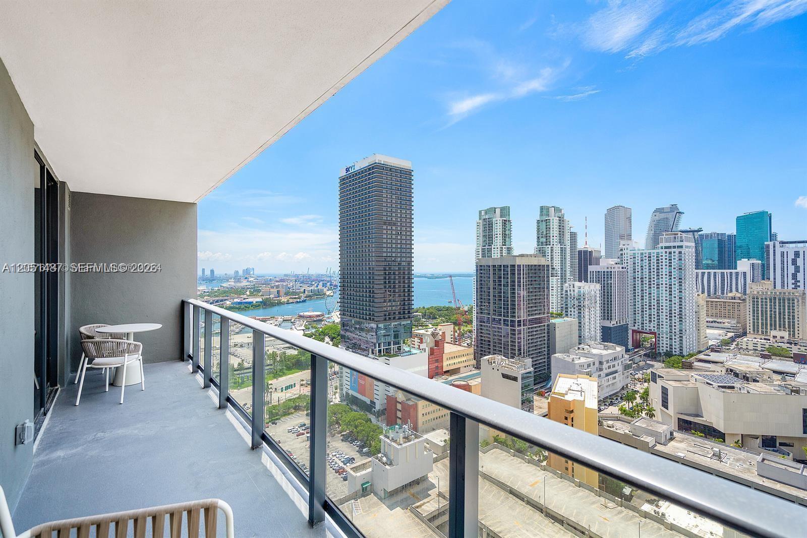 A balcony with a small table and chairs overlooks a dense city skyline and water — 159 NE 6th St # 2807, Miami FL 33132, MIAMI, FL
