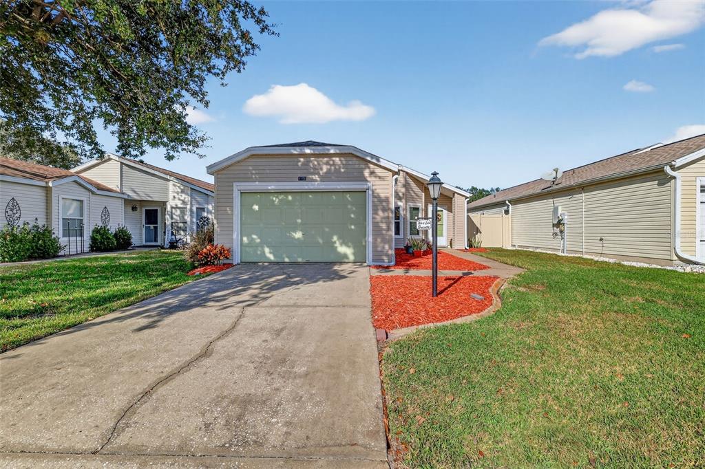 A single-story house with beige siding, a green garage door, and red mulch landscaping — 4900 HERON RUN CIRCLE, LEESBURG, FL