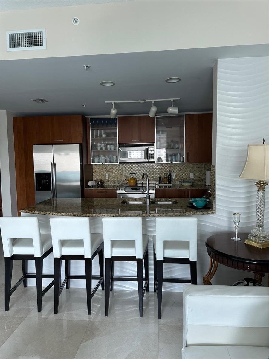 A kitchen features wood cabinets, granite countertops, stainless steel appliances, and four white bar stools — 1945 S Ocean Drive # 609, Hallandale Beach FL 33009, HALLANDALE BEACH, FL