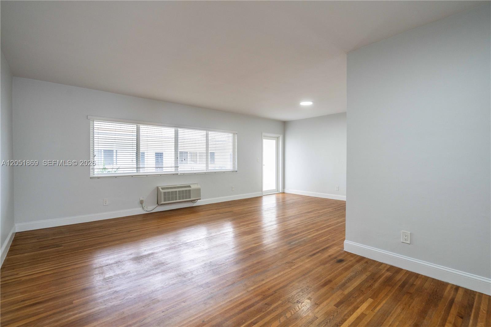 An empty room features hardwood floors, a large window with blinds, and a wall-mounted AC unit — 8330 Byron Ave # 7, Miami Beach FL 33141, MIAMI BEACH, FL