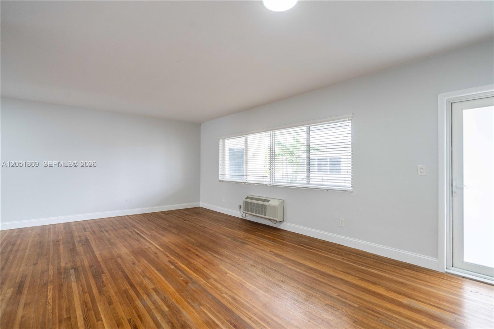 An empty room features hardwood floors, light gray walls, a window with blinds, and an AC unit — 8330 Byron Ave # 7, Miami Beach FL 33141, MIAMI BEACH, FL