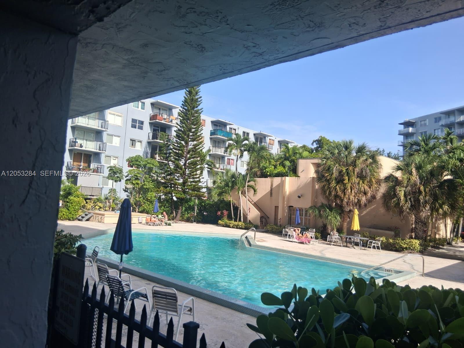 A swimming pool with lounge chairs and umbrellas is surrounded by apartment buildings and palm trees — 1470 NE 125th Ter # 812, North Miami FL 33161, NORTH MIAMI, FL