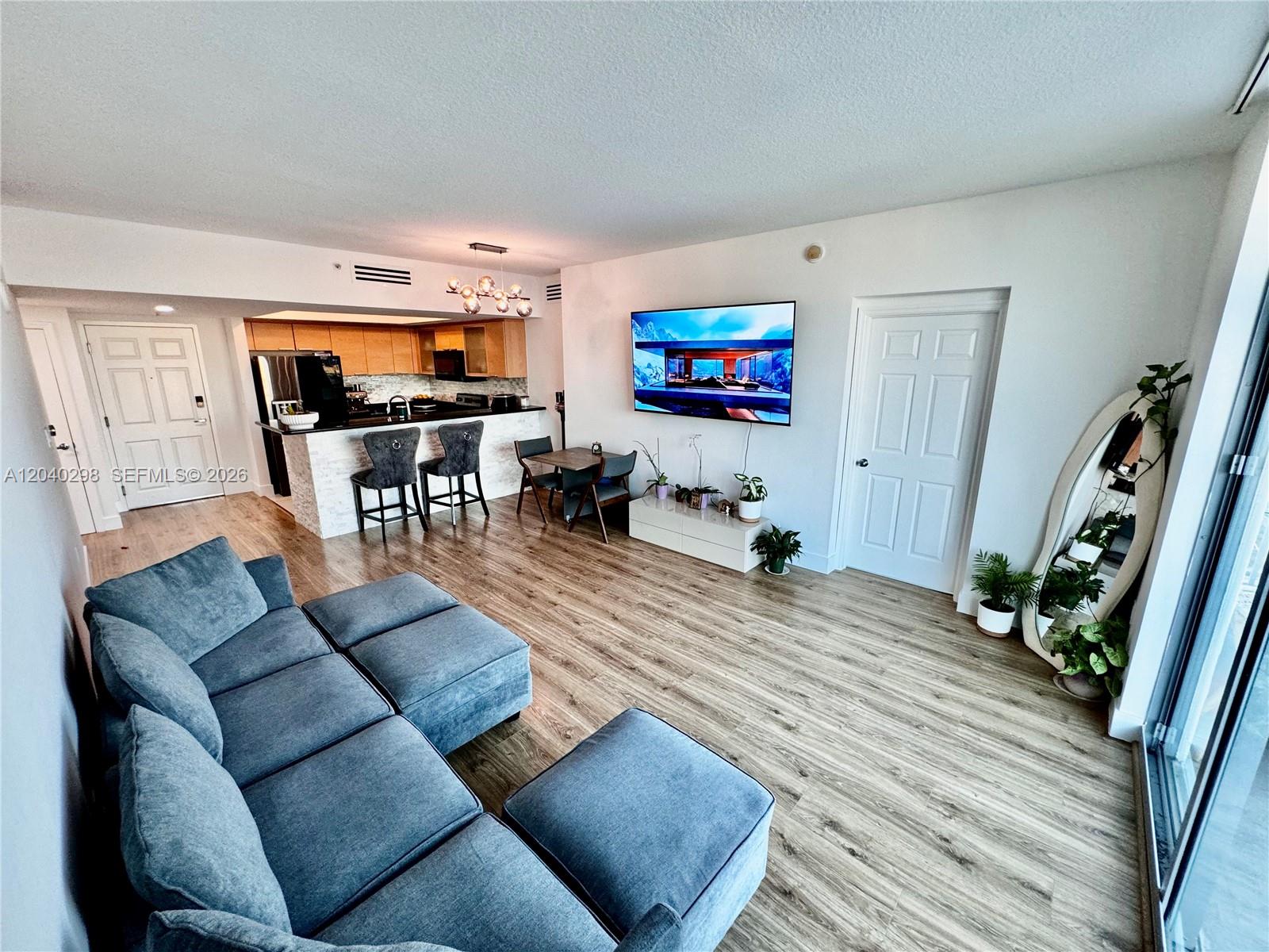An open-concept living room features a blue sectional sofa, a wall-mounted television, and a kitchen — 140 S Dixie Hwy # 921, Hollywood FL 33020, HOLLYWOOD, FL