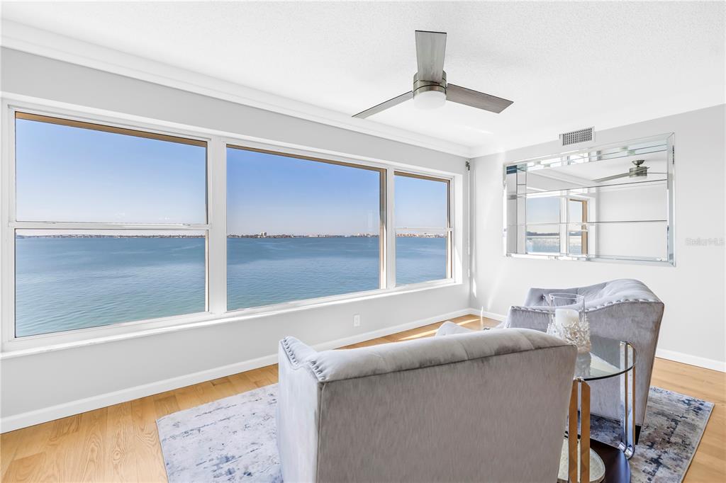 A living room features light wood floors, two gray armchairs, and large windows overlooking water — 6020 SHORE BOULEVARD S UNIT 712, GULFPORT, FL