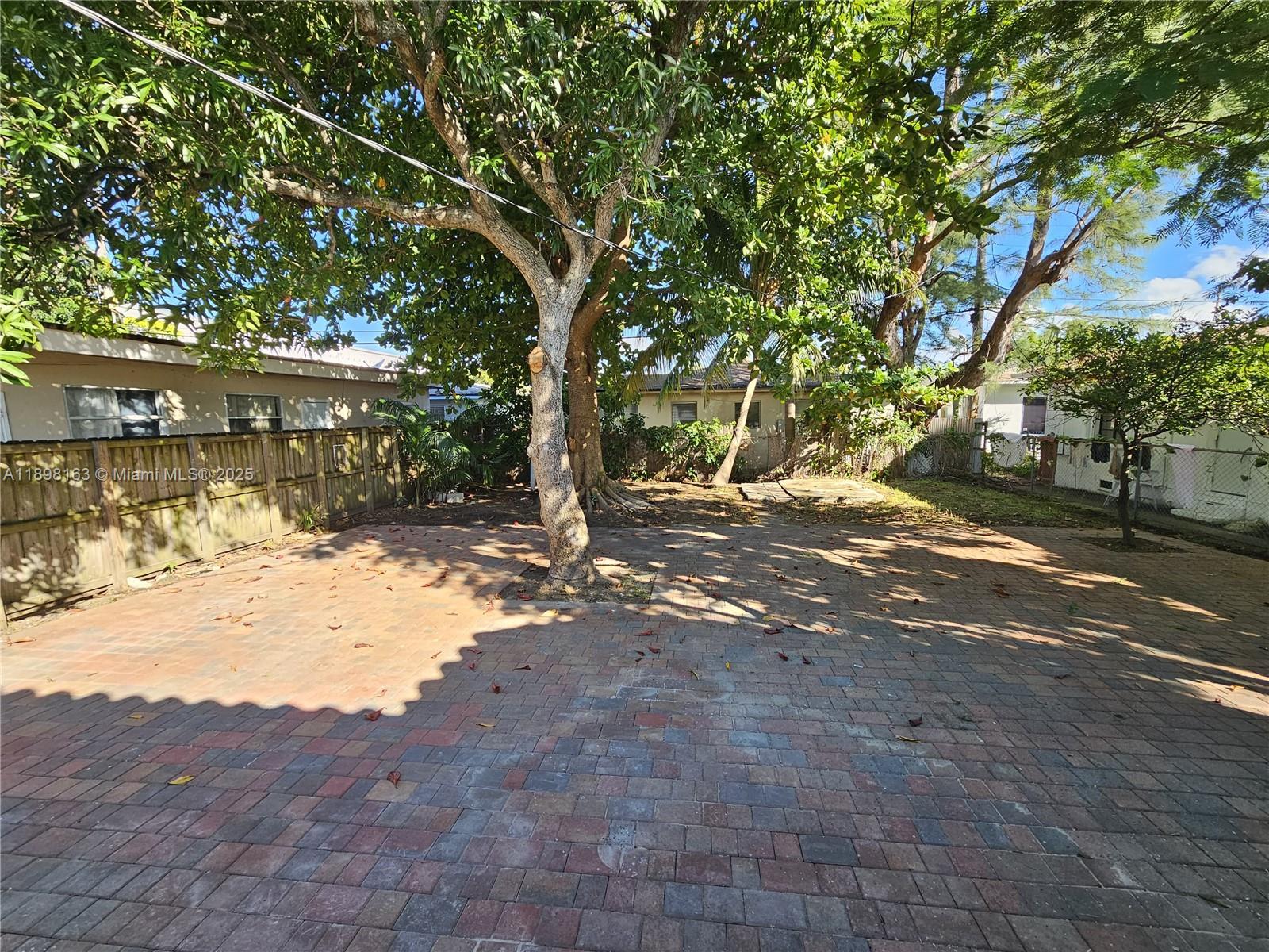 A paved backyard patio features a large central tree and a wooden fence — 1915 Normandy Drive, Miami Beach FL 33141, MIAMI BEACH, FL