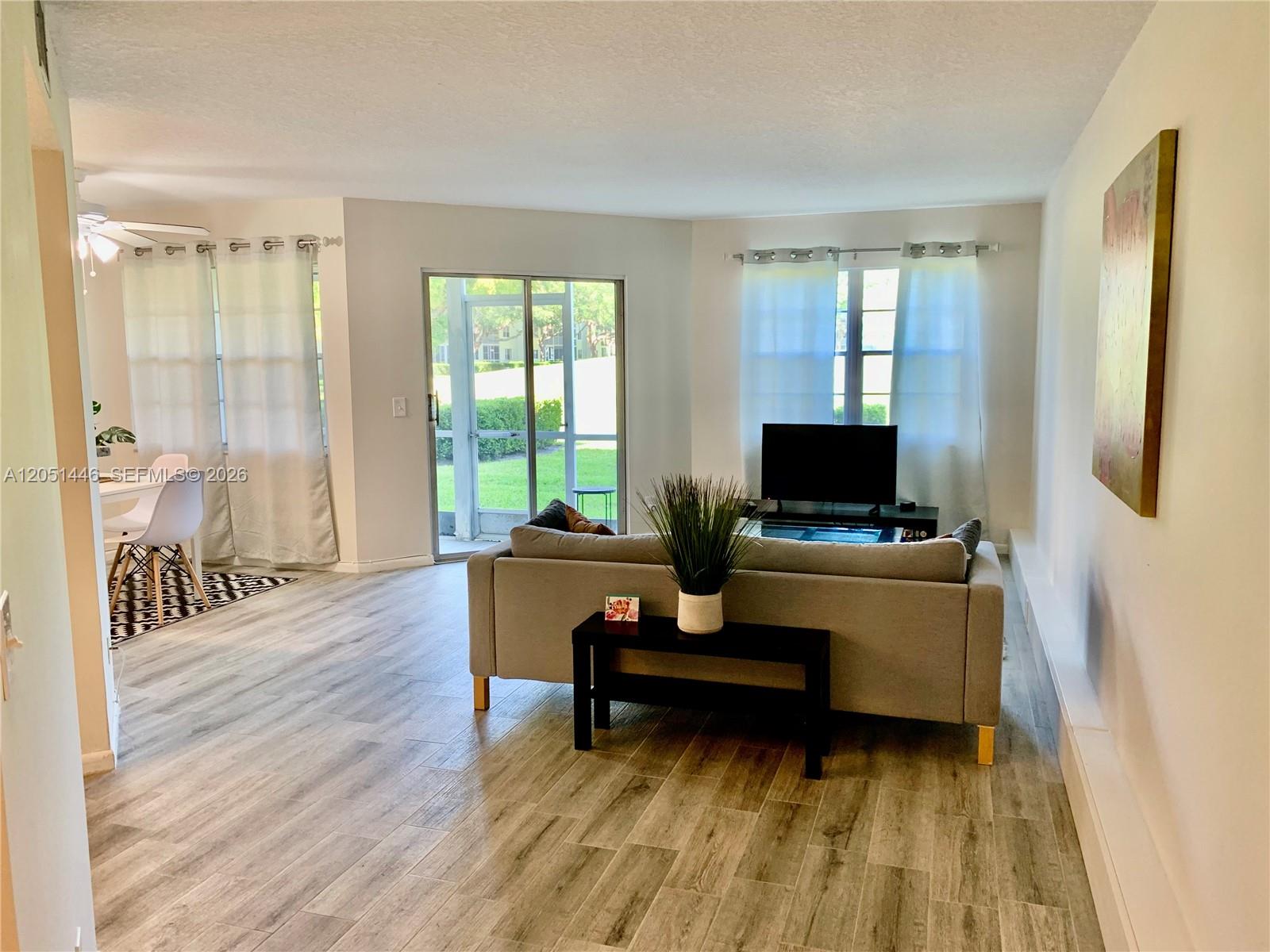 A living room with light wood-look flooring, a grey sofa, and a sliding glass door — PEMBROKE PINES, FL