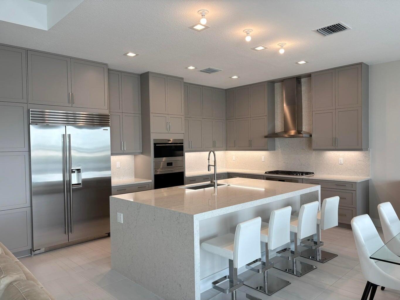 A modern kitchen features gray cabinetry, a large quartz island with seating, and stainless appliances — 9937 Migration Point, Palm Beach Gardens FL 33412, PALM BEACH GARDENS, FL