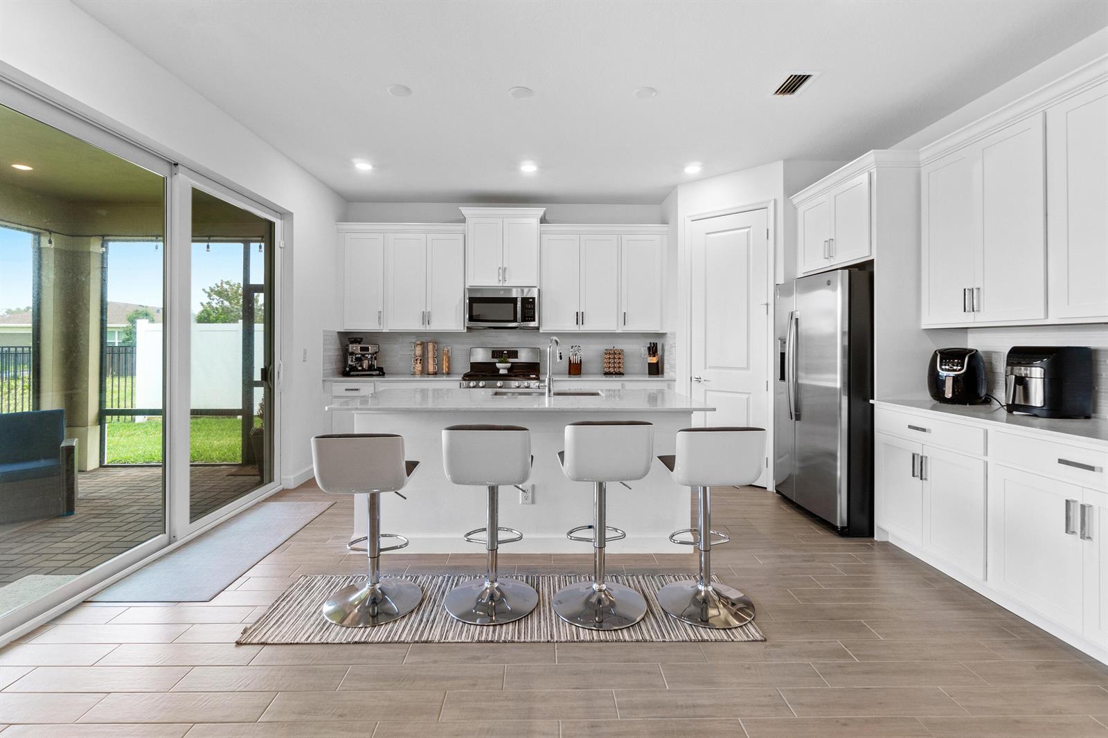 A modern kitchen features white cabinetry, a central island with four stools, and sliding doors — 1218 Timber Reap Trail, FL 33470, FL