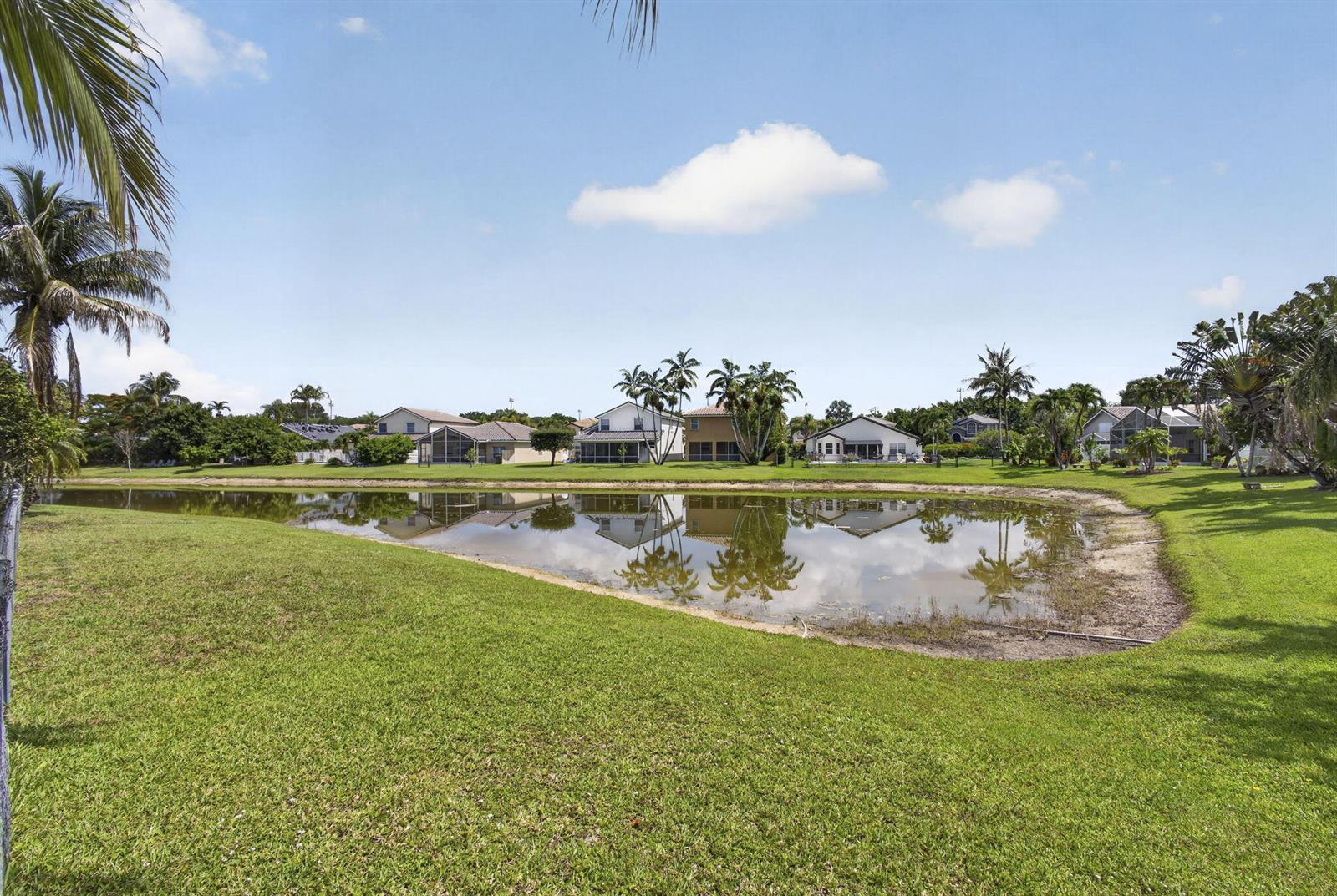 A grassy lawn slopes down to a pond reflecting a row of suburban houses — 11483 Beacon Pointe Lane, Wellington FL 33414, WELLINGTON, FL
