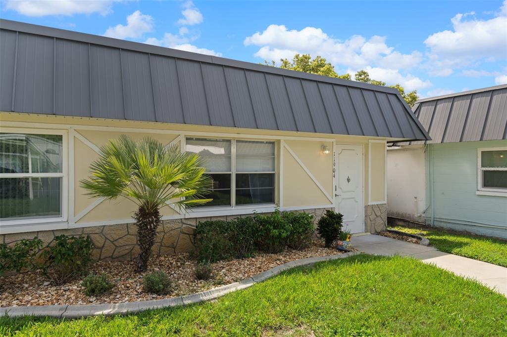 A single-story house exterior features a metal roof, light-colored siding, stone accents, and a palm tree — 11904 BAYONET LANE, NEW PORT RICHEY, FL