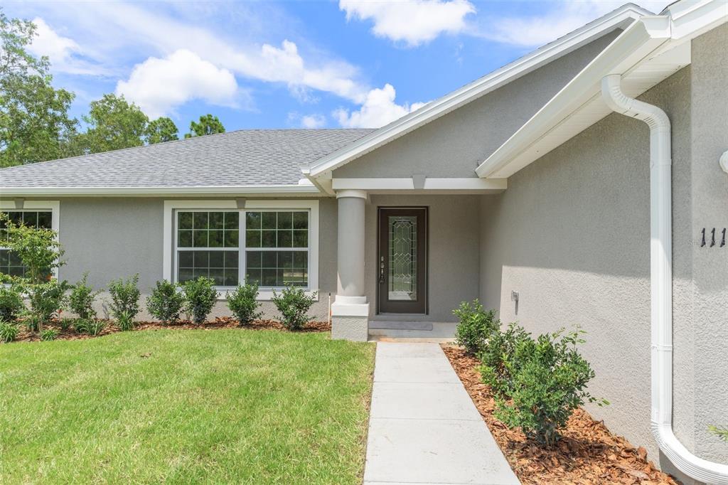A gray stucco house exterior features a front walkway, a glass-paneled door, and landscaping — 16375 KAKAWI ROAD, BROOKSVILLE, FL