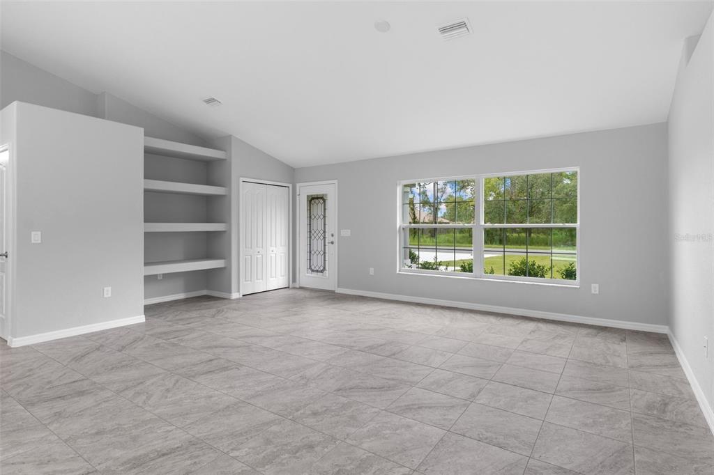 A bright, empty living room features gray tile flooring, built-in wall shelves, and a front door — 16375 KAKAWI ROAD, BROOKSVILLE, FL
