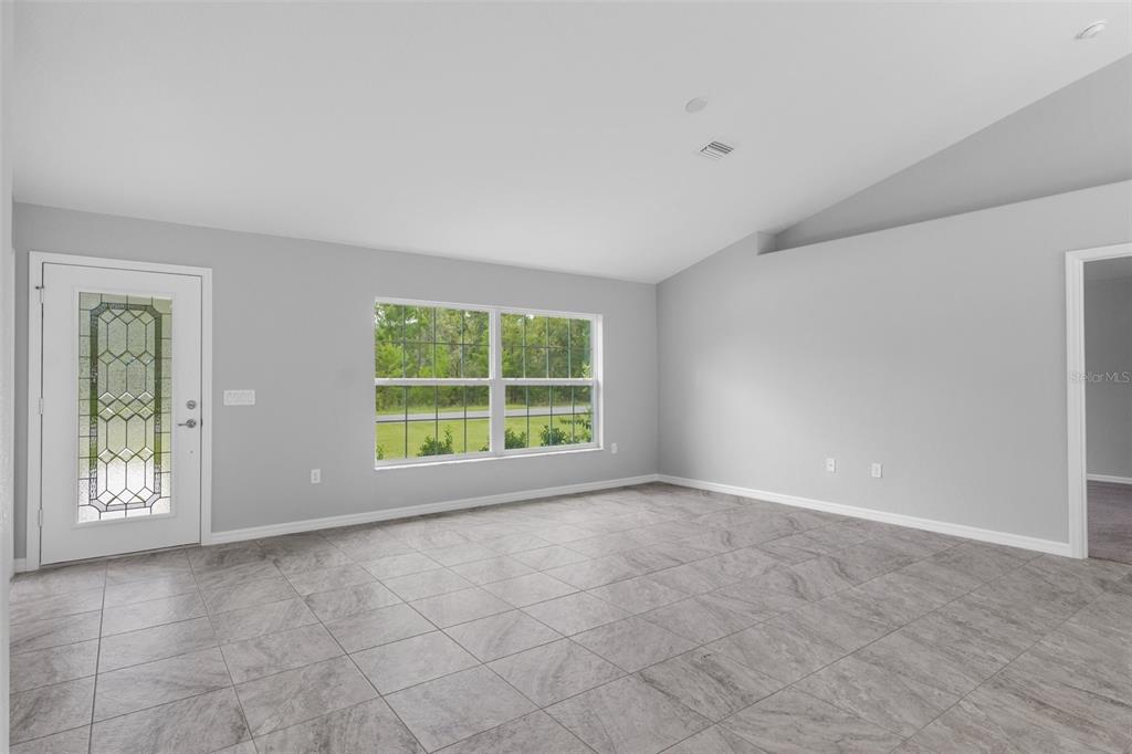 A bright, empty room features light gray walls, large windows, and gray tiled flooring — 16375 KAKAWI ROAD, BROOKSVILLE, FL