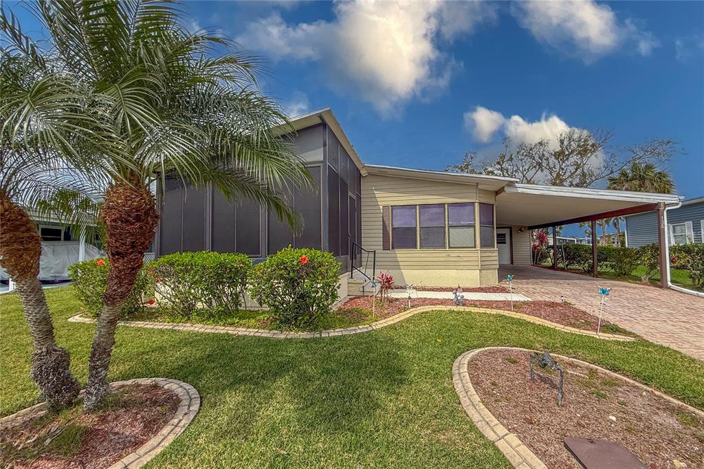 A single-story mobile home with a screened porch, carport, and landscaped yard under a blue sky — 135 HUMMINGBIRD AVENUE, ELLENTON, FL