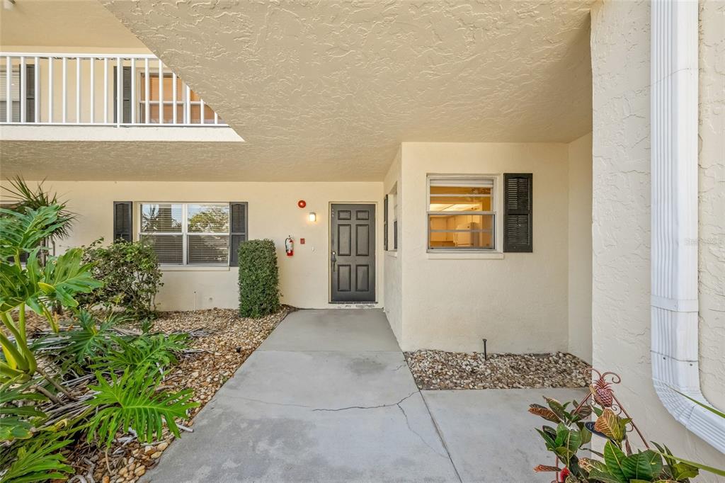 A concrete walkway leads to the dark front door of a beige stucco building — 7860 PALM AIRE LANE UNIT 105, SARASOTA, FL