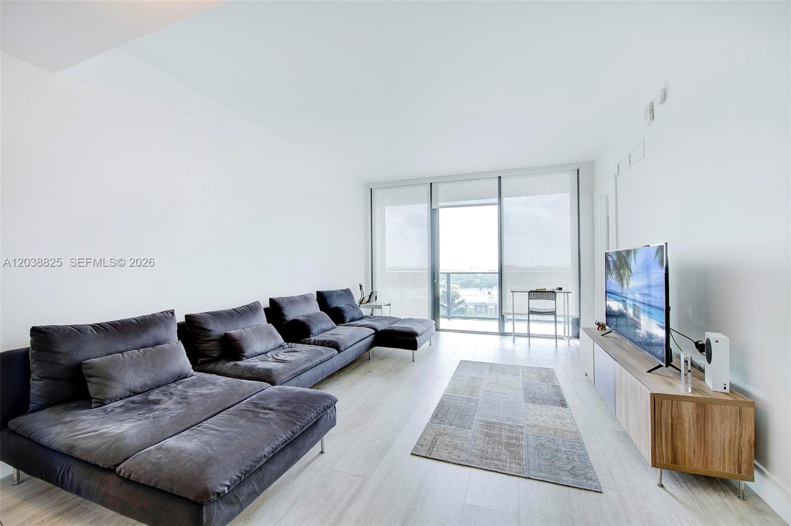 A modern living room features a large gray sectional sofa, a television, and a balcony — 121 NE 34th St # 1111, Miami FL 33137, MIAMI, FL