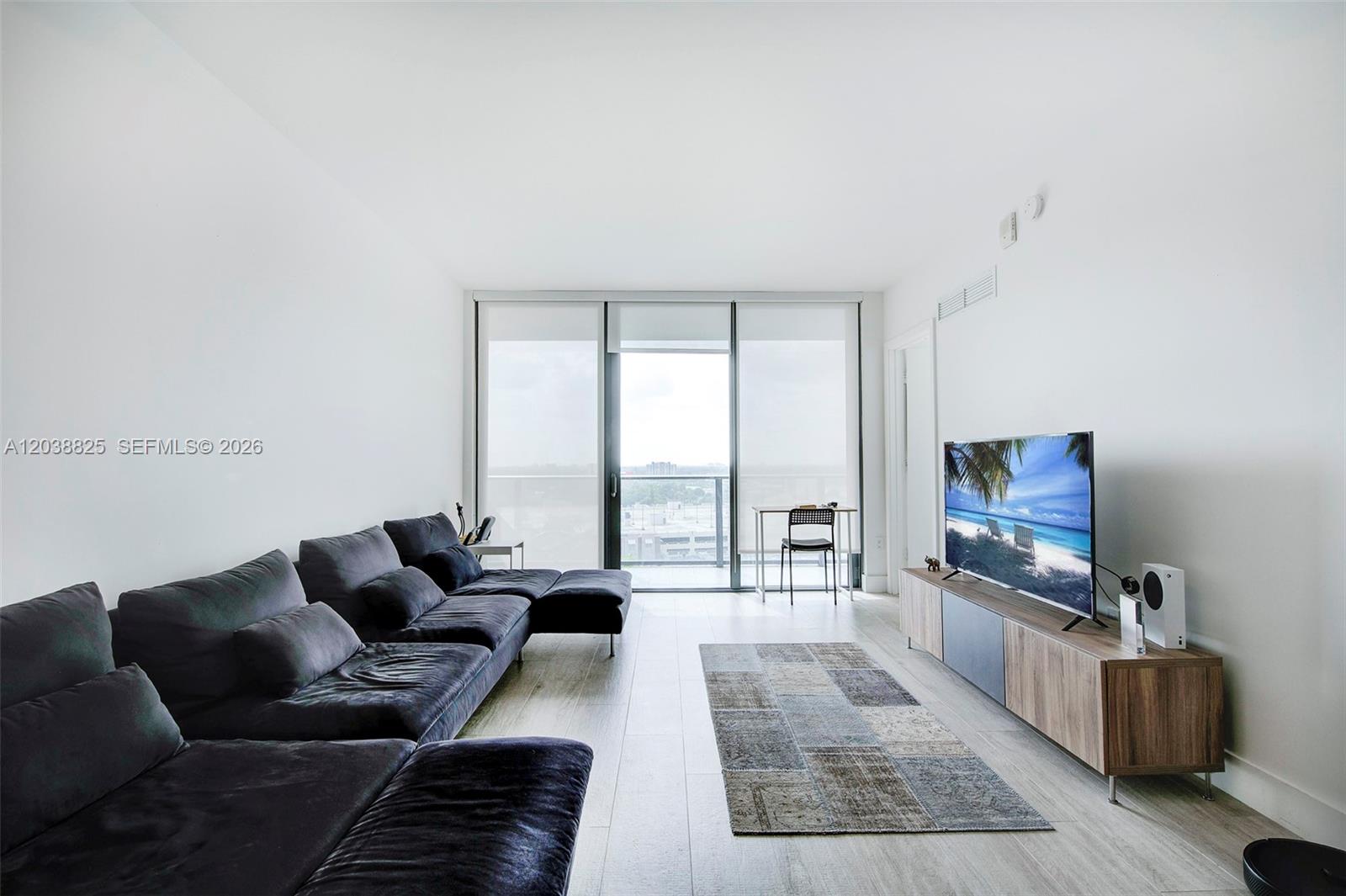 A modern living room features a dark sectional sofa, wood media console, and floor-to-ceiling windows — 121 NE 34th St # 1111, Miami FL 33137, MIAMI, FL