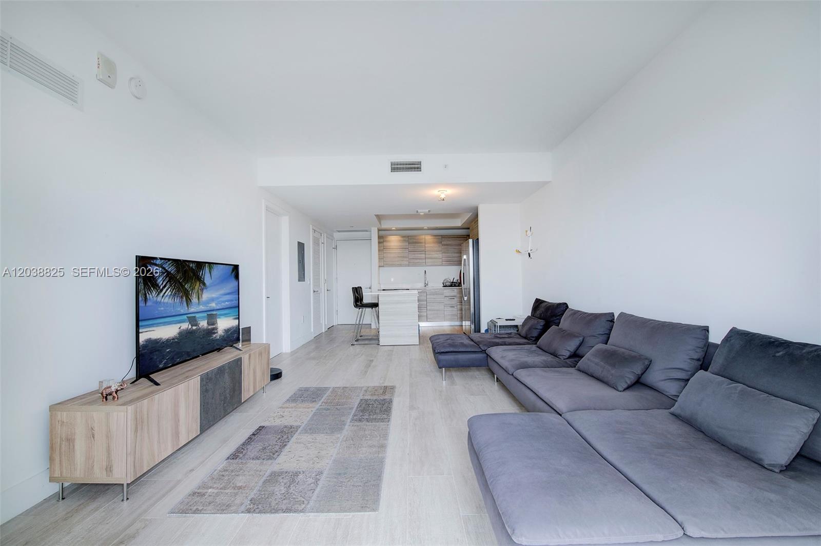 A modern living room features a grey sectional sofa, a television console, and an open kitchen — 121 NE 34th St # 1111, Miami FL 33137, MIAMI, FL