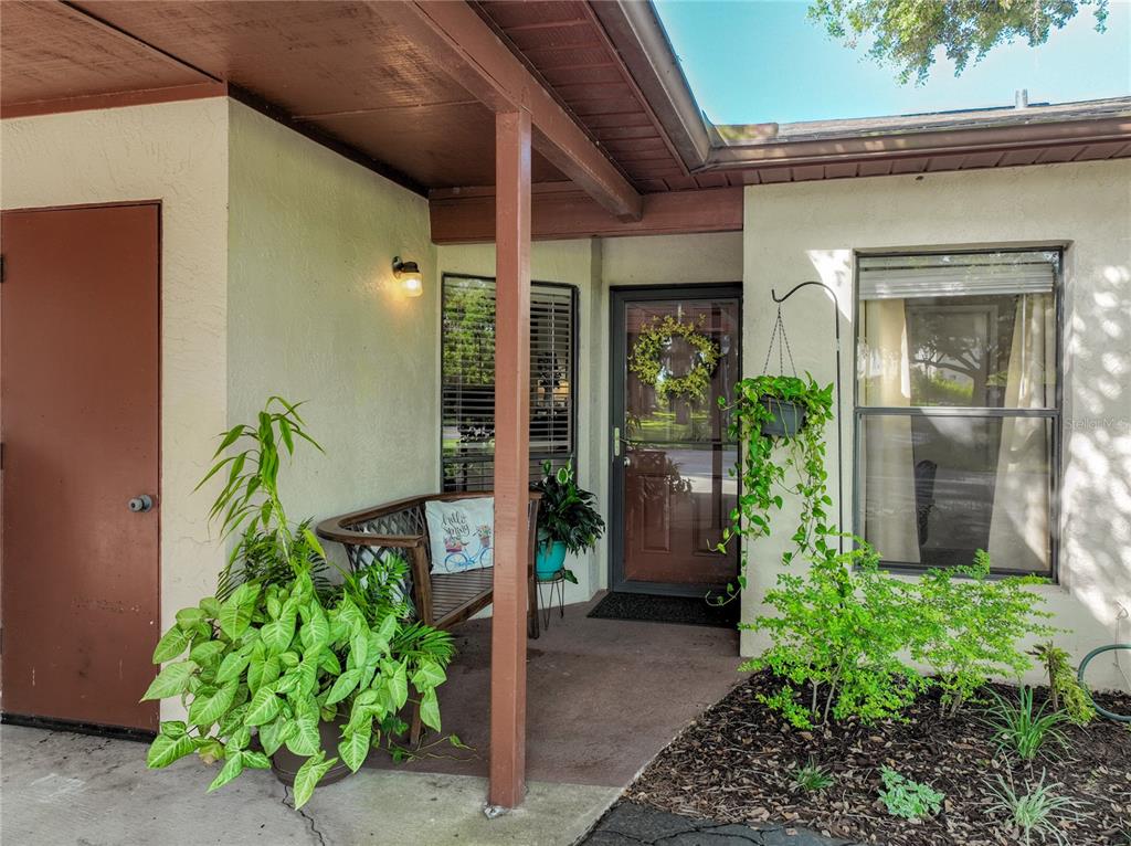 A covered entryway features a brown front door, a wooden bench, and several potted plants — 4300 BAYWOOD BOULEVARD UNIT C106, MOUNT DORA, FL