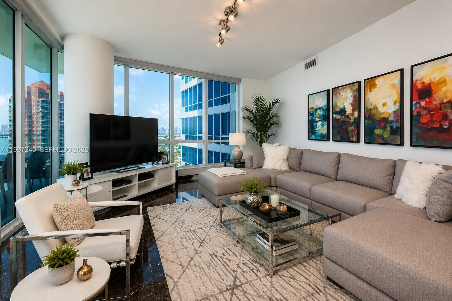 A modern living room features a grey sectional sofa, glass coffee table, and floor-to-ceiling windows — 100 S Pointe Dr # 3101, Miami Beach FL 33139, MIAMI BEACH, FL
