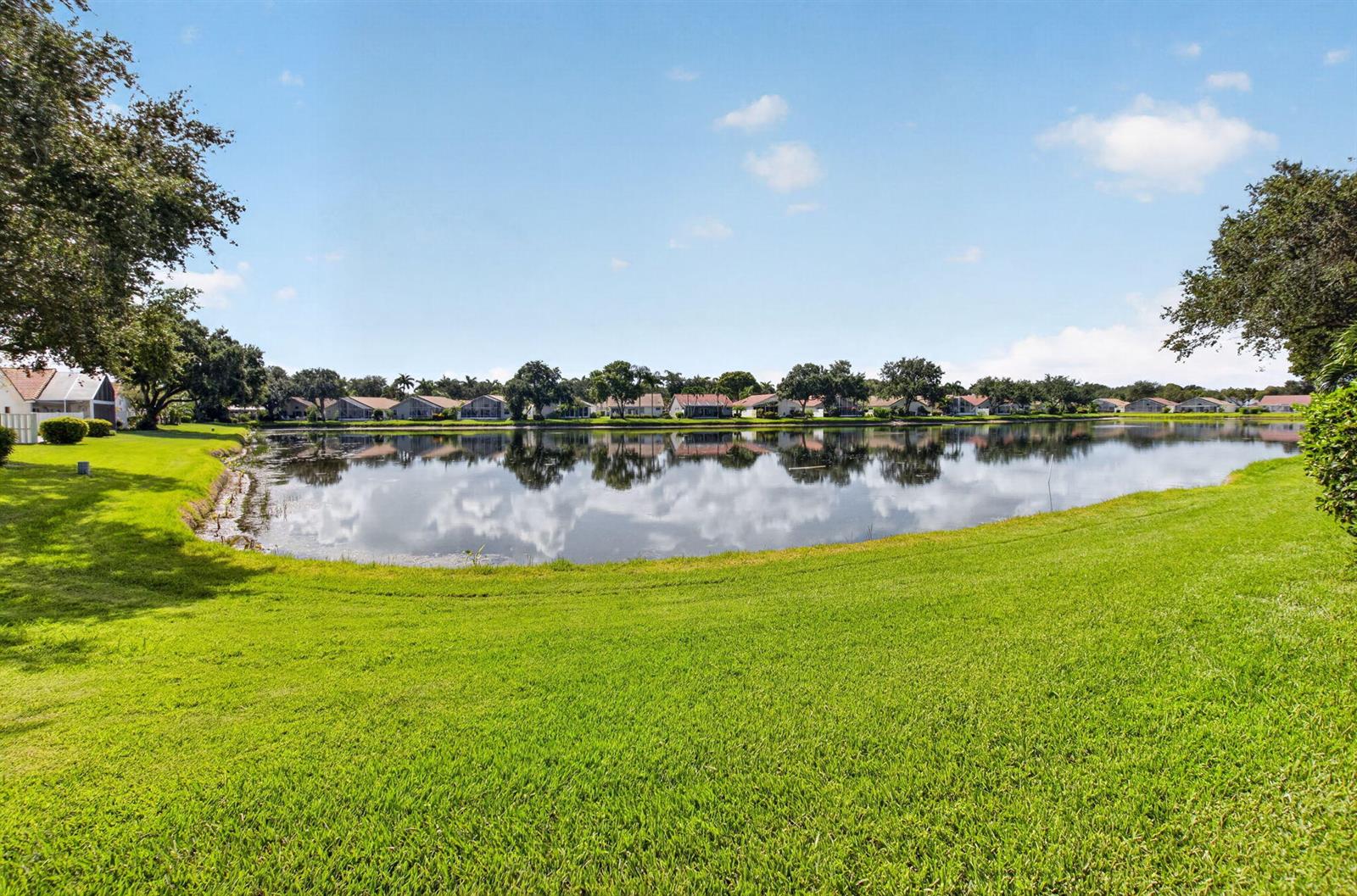A grassy lawn overlooks a calm lake with houses visible along the distant shoreline — 9875 Harbour Lake Circle, Boynton Beach FL 33437, BOYNTON BEACH, FL