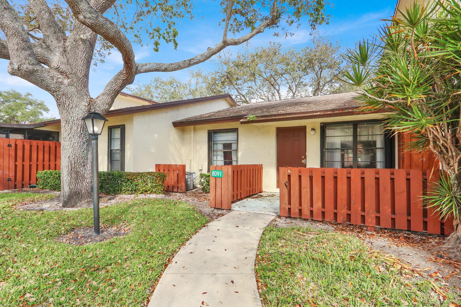 A single-story residential unit with a brown door, wooden fence, and a large oak tree — 8091 SE Villa Circle, Hobe Sound FL 33455, HOBE SOUND, FL