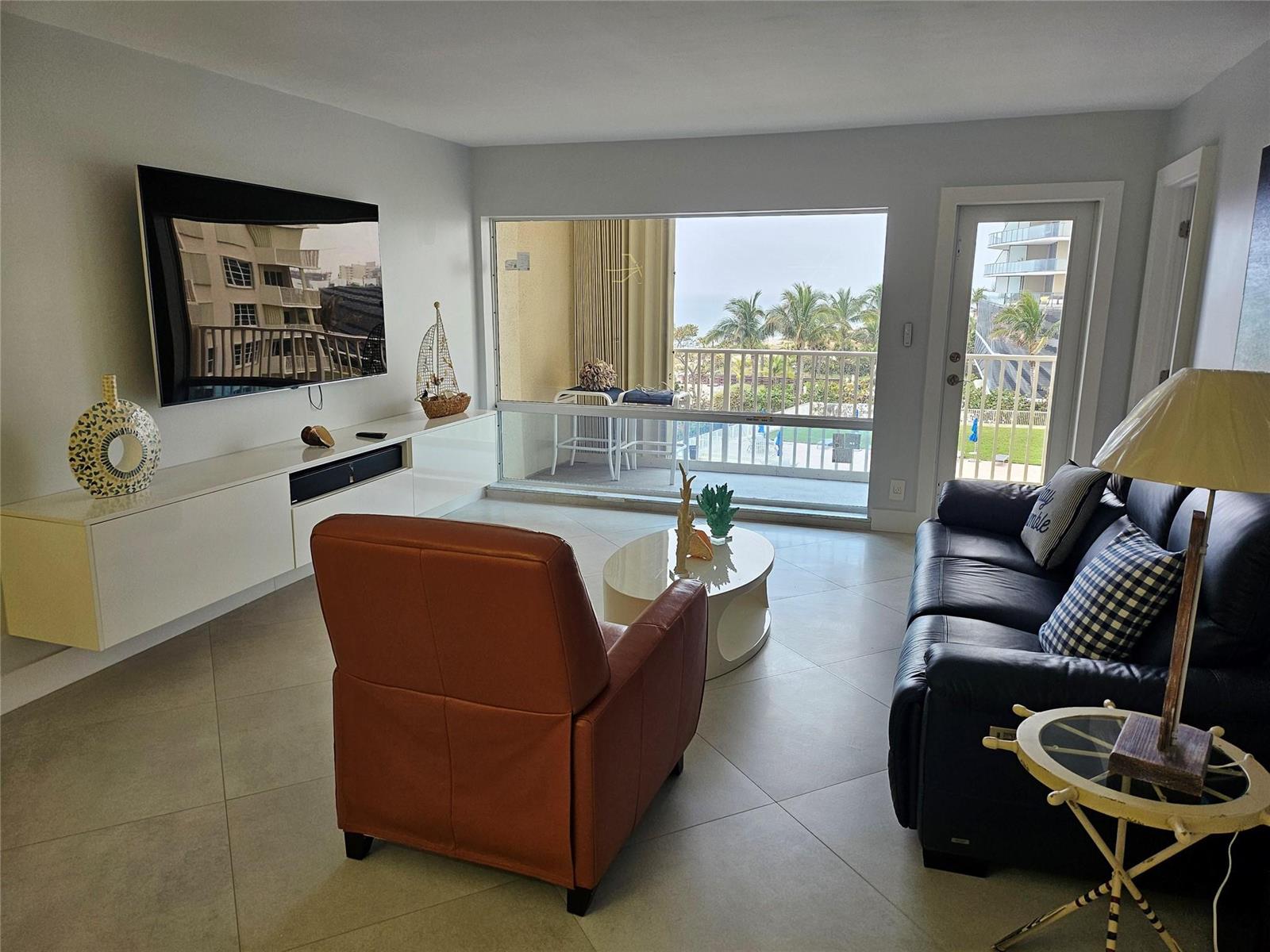 A living room with a leather sofa, recliner, media console, and a balcony overlooking palm trees — 750 N Ocean Boulevard # 503, Pompano Beach FL 33062, POMPANO BEACH, FL