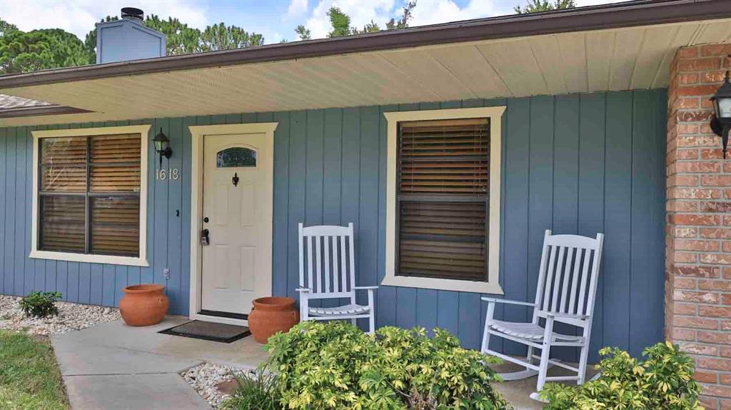 A blue house exterior features a white front door, two rocking chairs, and brick accents — 1618 WILLOW OAK DRIVE, EDGEWATER, FL