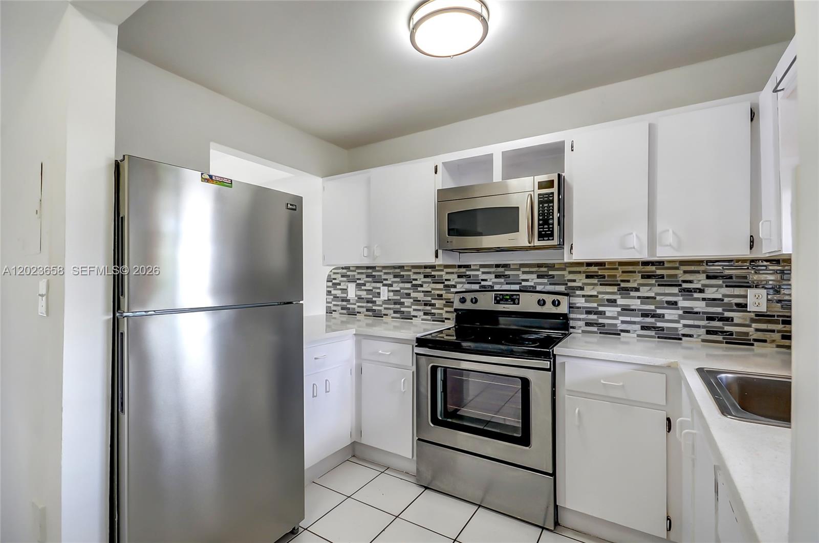 A kitchen features white cabinets, stainless steel appliances, a tiled backsplash, and white floor tiles — 3021 W 76th St # 203, Hialeah FL 33018, HIALEAH, FL