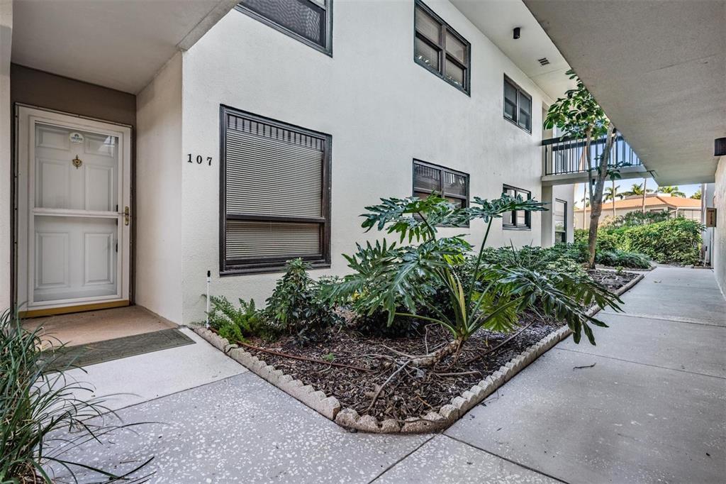 A white apartment building exterior features a numbered front door and a landscaped garden bed — 6294 BAHIA DEL MAR CIRCLE UNIT 107, ST PETERSBURG, FL