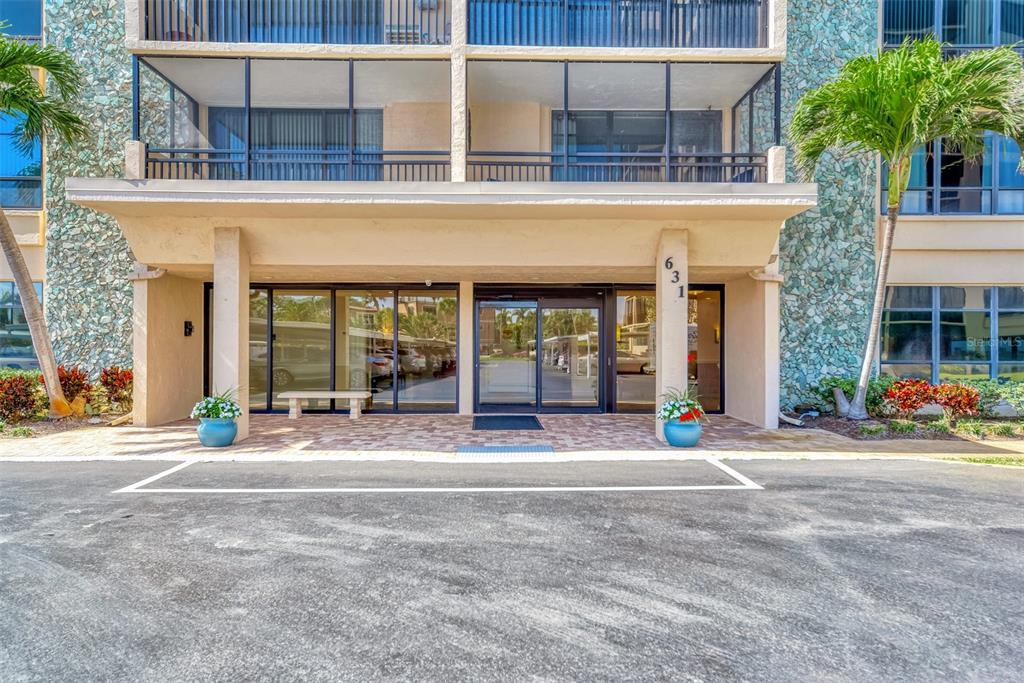 The exterior entrance of a multi-story building features glass doors, stone siding, and two palm trees — 631 ALHAMBRA ROAD UNIT 1004, VENICE, FL