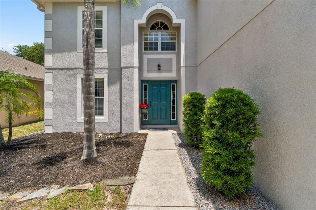 A concrete walkway leads to a teal front door of a two-story stucco house — 3216 BELLERICAY LANE, LAND O LAKES, FL