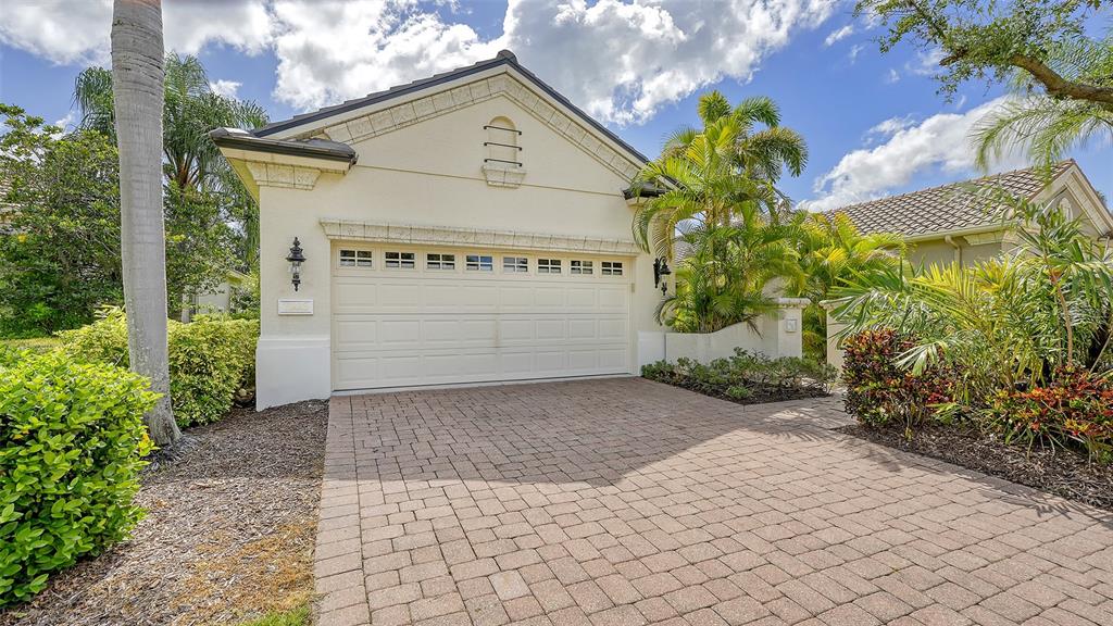 A beige house with a two-car garage and a paved driveway surrounded by tropical landscaping — 7223 PRESIDIO GLEN, LAKEWOOD RANCH, FL