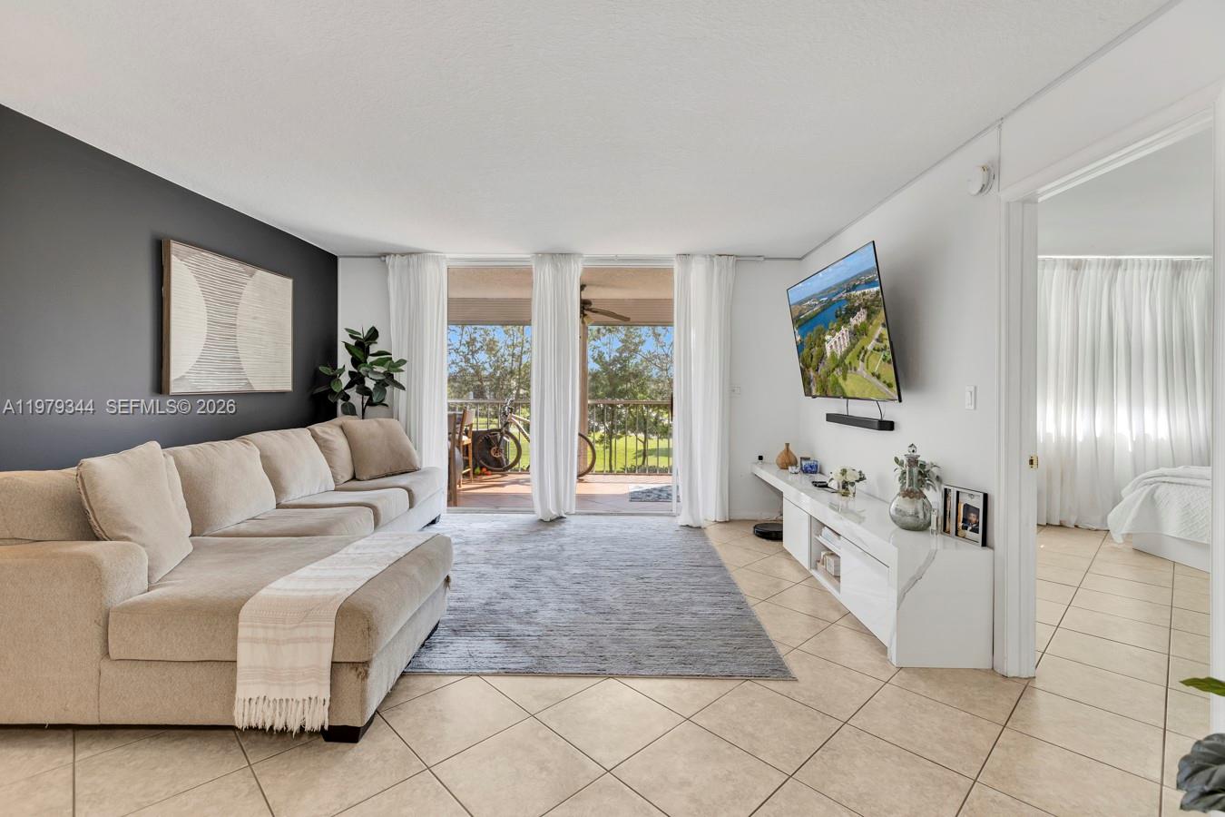 A living room features a beige sectional sofa, a wall-mounted television, and a balcony view — 4550 NW 18th Ave # 206, Deerfield Beach FL 33064, DEERFIELD BEACH, FL