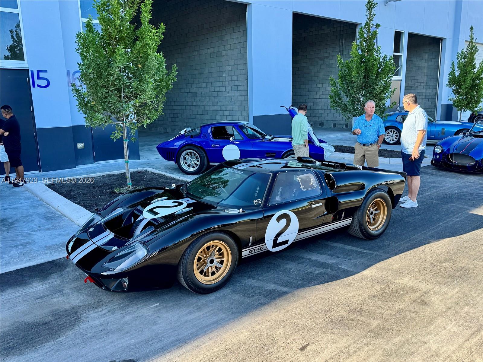 A black Ford GT40 race car is parked outside a building with other classic cars — 15375 Corporate Rd # 17, Jupiter FL 33478, JUPITER, FL