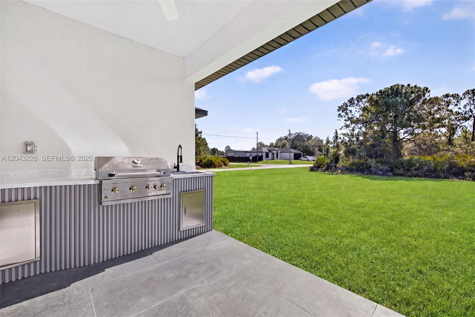 An outdoor kitchen with a stainless steel grill sits on a covered patio overlooking grass — 3706 N Gene Ave, Lehigh Acres FL 33971, LEHIGH ACRES, FL