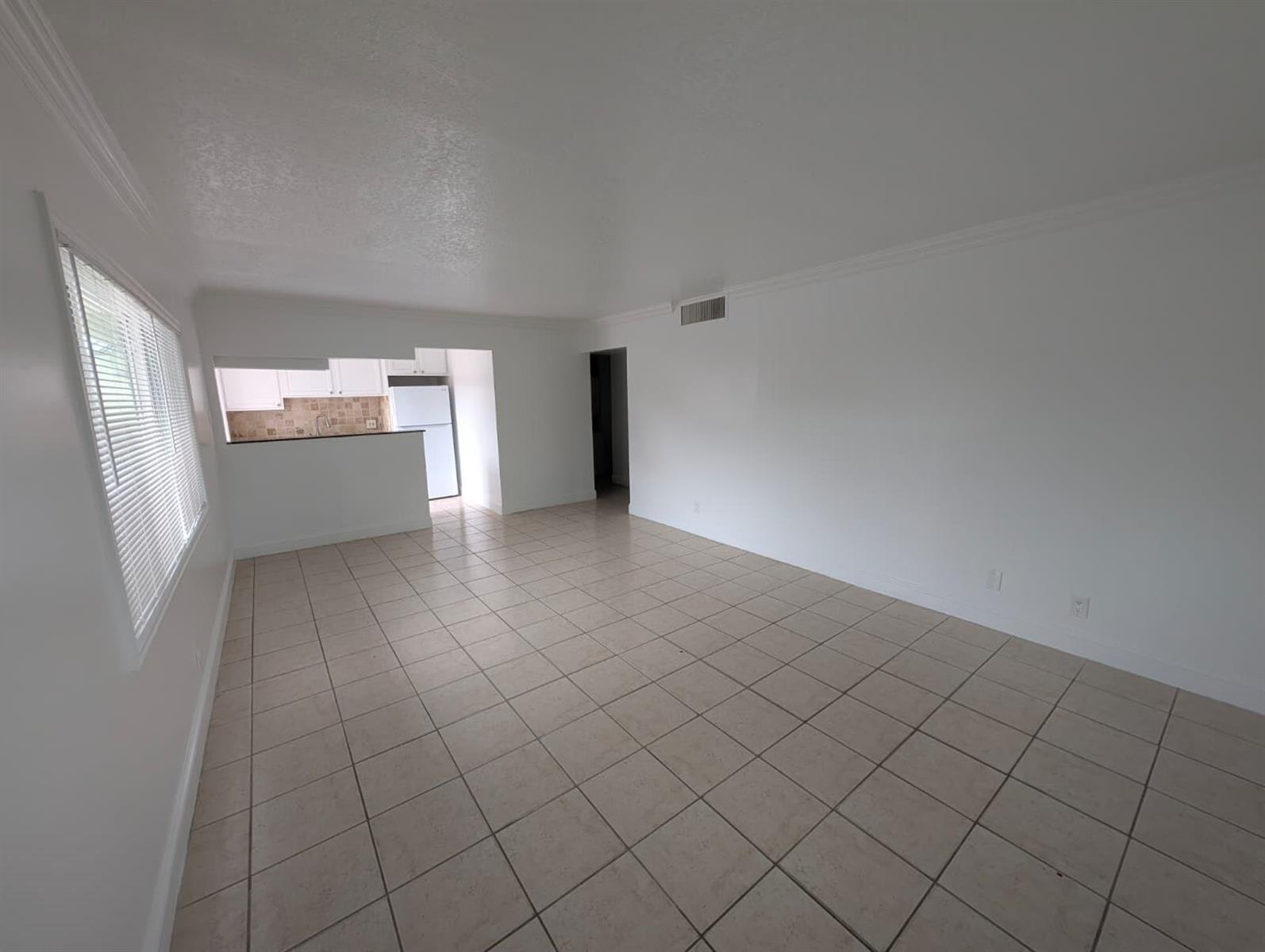 An empty living room with light-colored tile flooring and white walls leads to a kitchen — 817 Lighthouse Drive Apt B, North Palm Beach FL 33408, NORTH PALM BEACH, FL