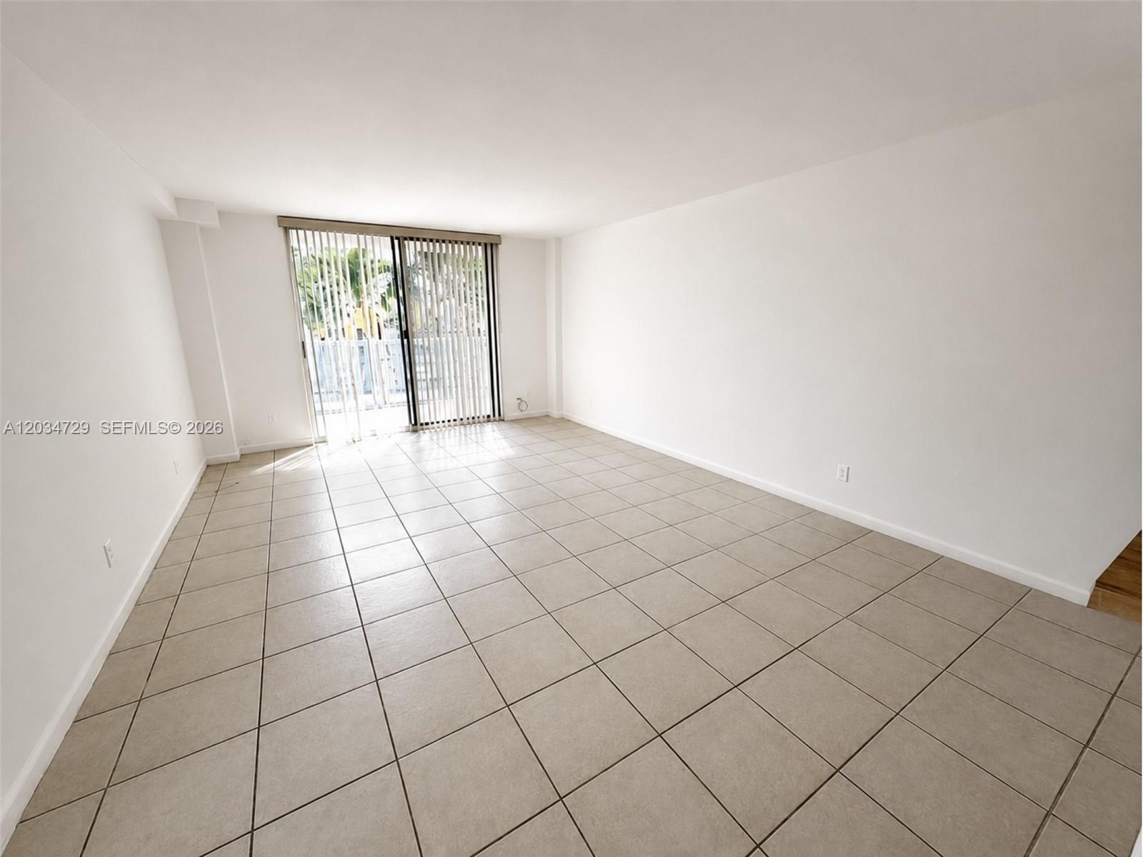 An empty living room features white walls, beige tile flooring, and sliding glass balcony doors — 13499 Biscayne Blvd # 504, North Miami FL 33181, NORTH MIAMI, FL