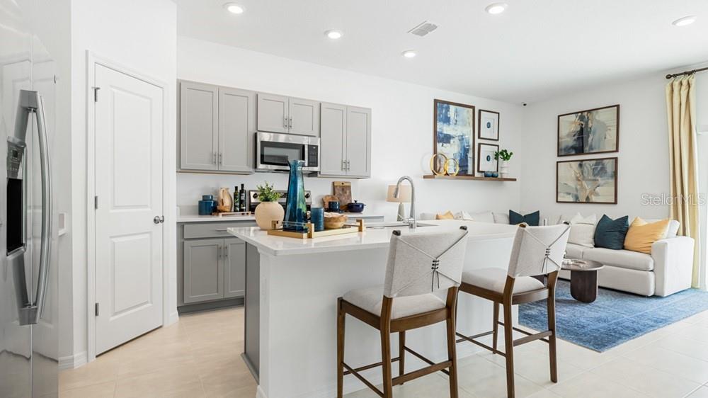 A modern kitchen features gray cabinets, a white island with bar stools, and an adjacent living area — 7117 PINELIT COVE, NOKOMIS, FL