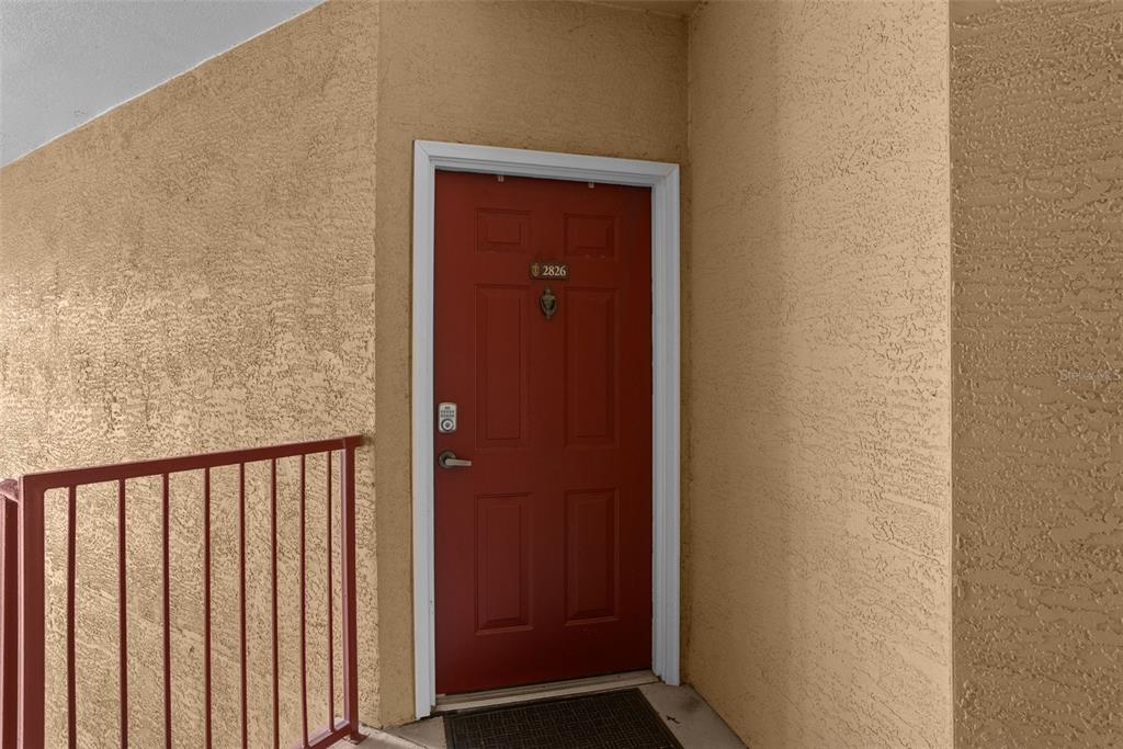 A red front door with a digital lock is set into a textured tan wall — 2141 PORTOFINO PLACE UNIT 8-2826, PALM HARBOR, FL