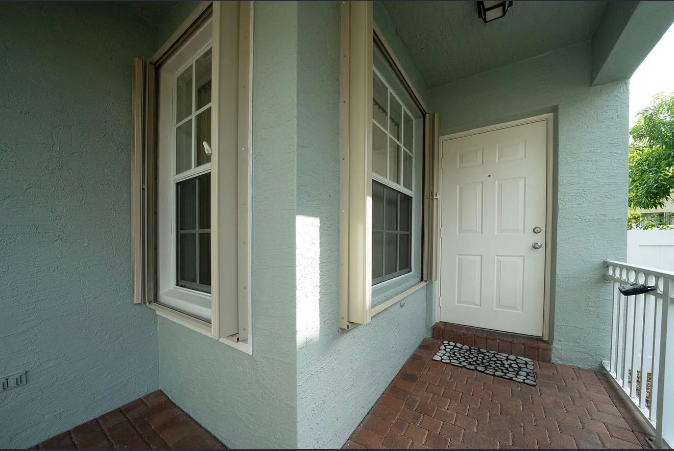 A covered porch features light green stucco walls, brick flooring, and a white entry door — 5144 Aurora Lake Circle, FL 33463, FL