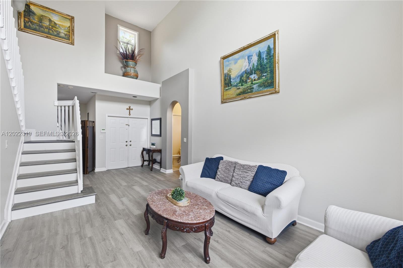 A living room features high ceilings, gray wood-look flooring, a white sofa, and a staircase — 16928 SW 35th St, Miramar FL 33027, MIRAMAR, FL