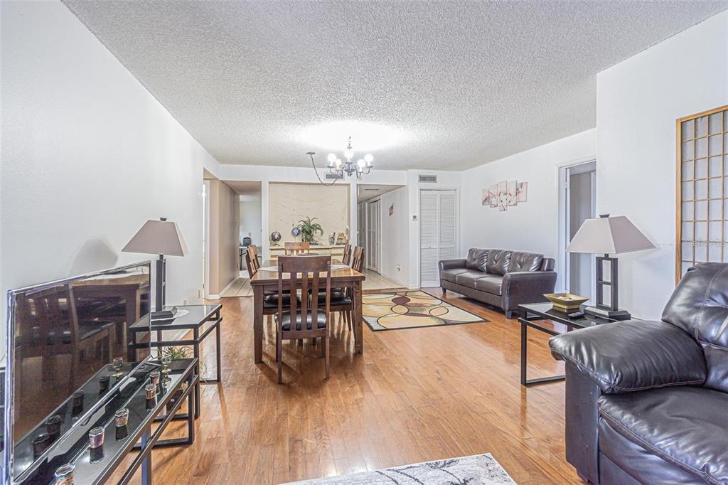 An open-concept living and dining area features hardwood floors, dark leather furniture, and a chandelier — 14820 RUE DE BAYONNE UNIT 301, CLEARWATER, FL