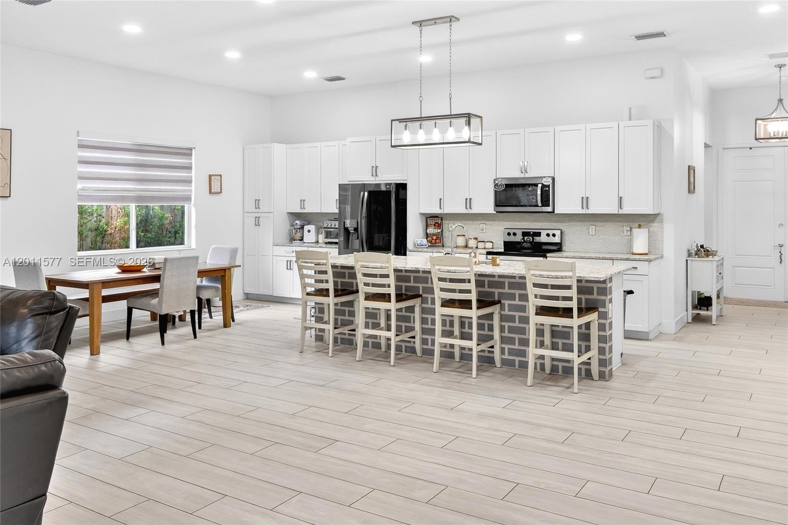 A bright, open-concept kitchen features white cabinetry, a brick-faced island, and light wood-look tile flooring — 15012 SW 177th Ter, Miami FL 33187, MIAMI, FL
