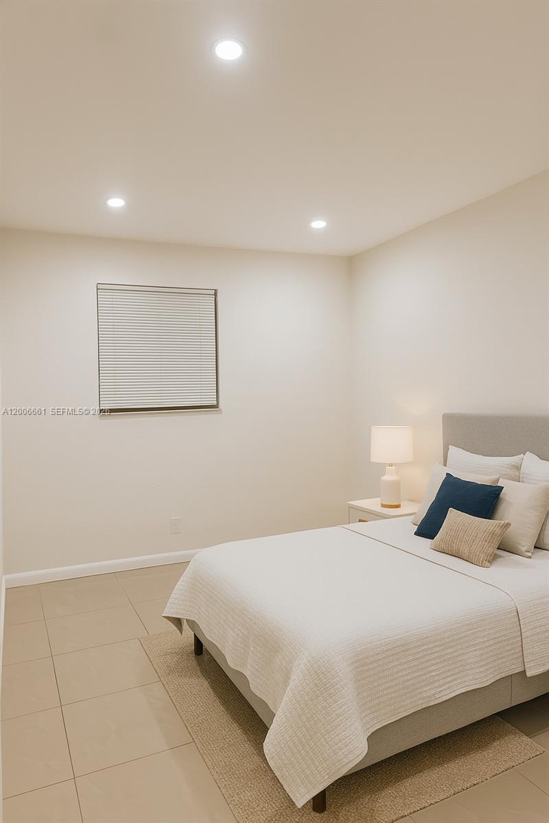 A bedroom features a bed with neutral bedding, a nightstand with a lamp, and tile flooring — 1046 Foster Rd # 0, Hallandale Beach FL 33009, HALLANDALE BEACH, FL