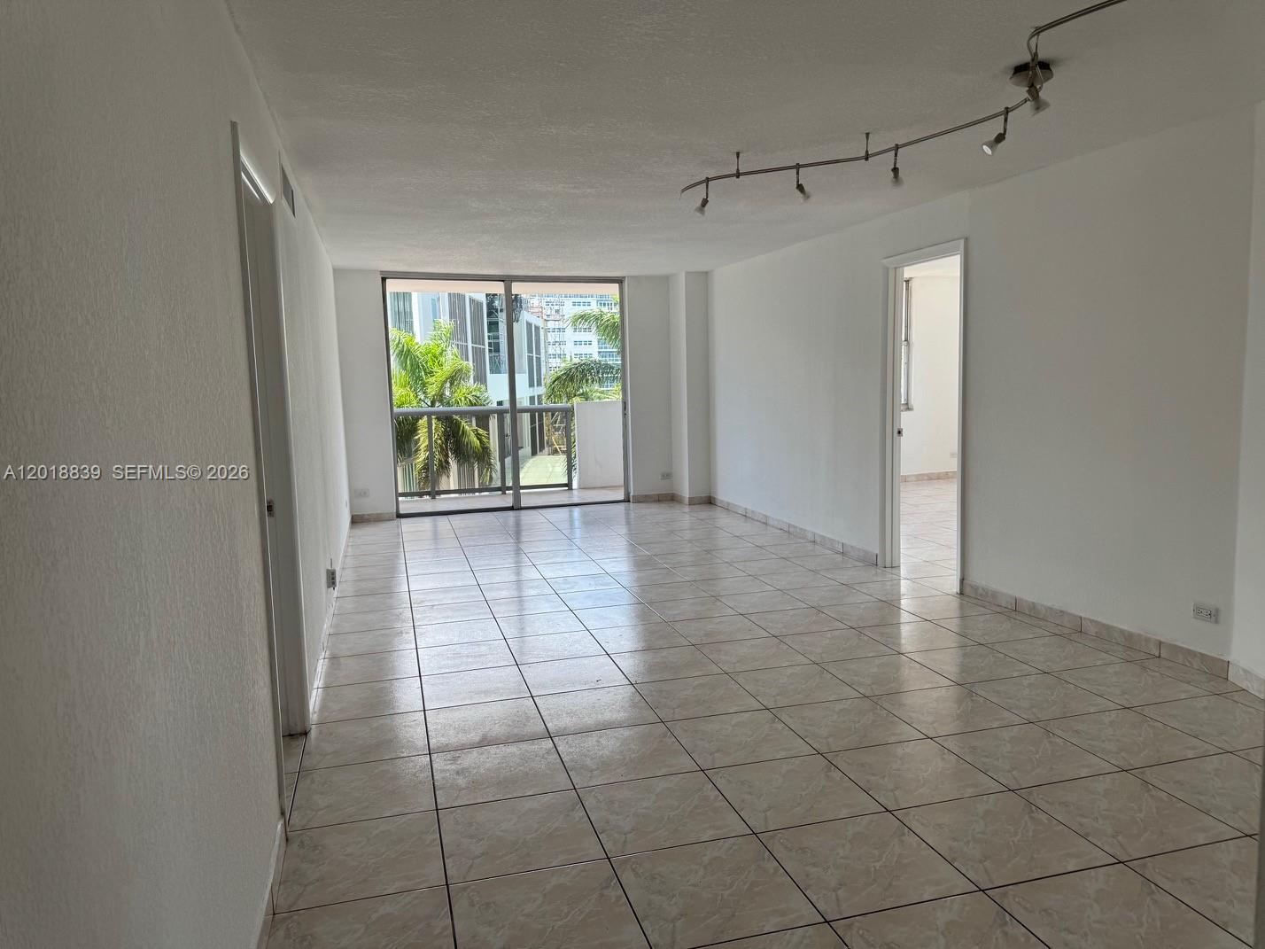 An empty living room features light-colored tile flooring, white walls, and a sliding glass balcony door — 6039 COLLINS AV # 537, Miami Beach FL 33140, MIAMI BEACH, FL