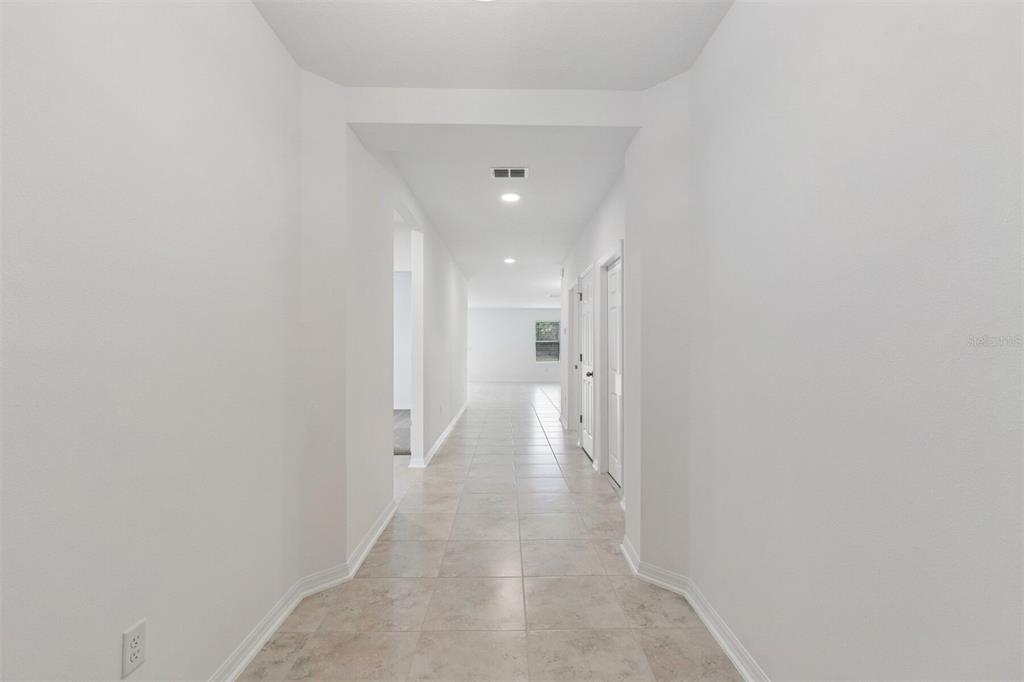 A long, white-walled hallway with light-colored tile flooring leads toward a bright, open living area — 8437 FOX DEN COVE, LAKELAND, FL