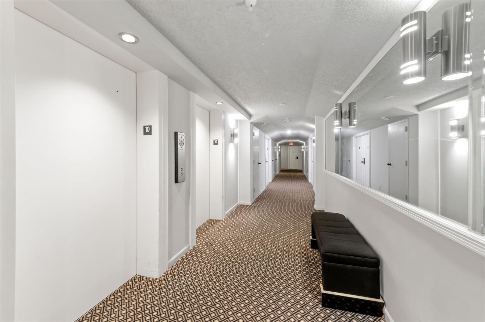 A long apartment hallway features patterned carpet, white walls, a bench, and mirrored wall accents — 1900 Consulate Place # 1004, West Palm Beach FL 33401, WEST PALM BEACH, FL